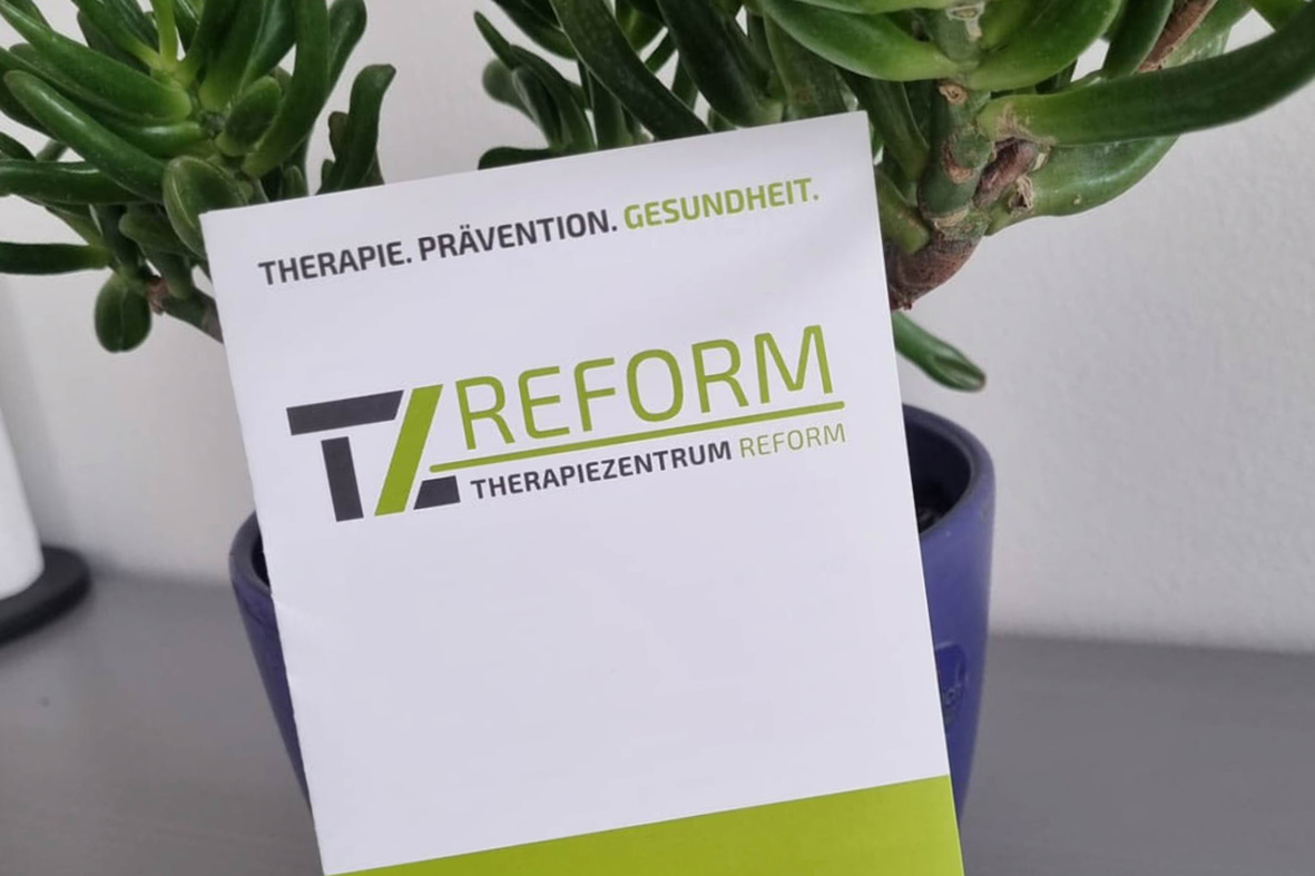 Modernes weißes Gebäude des Therapiezentrums Reform Magdeburg mit grünen Schildern, großen Fenstern und mit Sträuchern gesäumten Gehwegen.