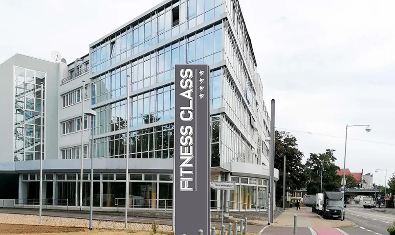 Modernes weißes Gebäude des Therapiezentrums Reform Magdeburg mit grünen Schildern, großen Fenstern und mit Sträuchern gesäumten Gehwegen.